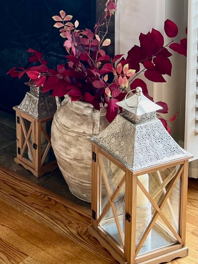 Two wooden lanterns with metal tops sit on a hardwood floor next to a large rustic vase filled with red leafy branches, creating a cozy simple fall home tour vibe in front of a dark fireplace and white wall.