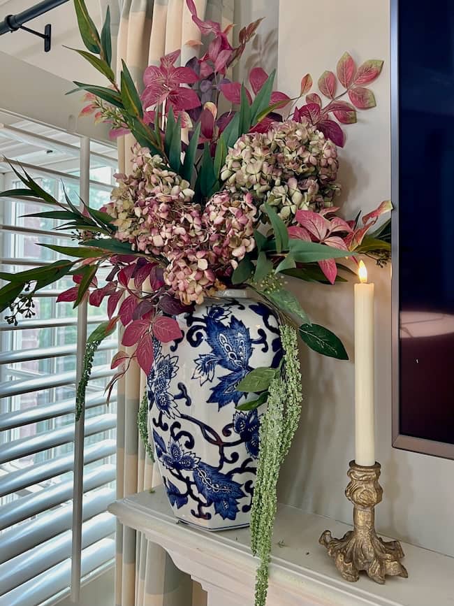 A large blue and white vase with floral patterns holds an arrangement of pink and green flowers and foliage. Beside it, a lit white candle in an ornate gold holder warms the mantle—perfect for a cozy simple fall home tour.