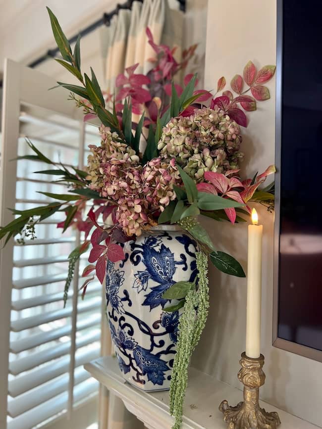 A blue and white ceramic vase with floral patterns holds a bouquet of pink hydrangeas, leafy branches, and greenery. Next to it, a lit white candle sits in a gold holder on the mantelpiece—a cozy touch for a simple fall home tour.