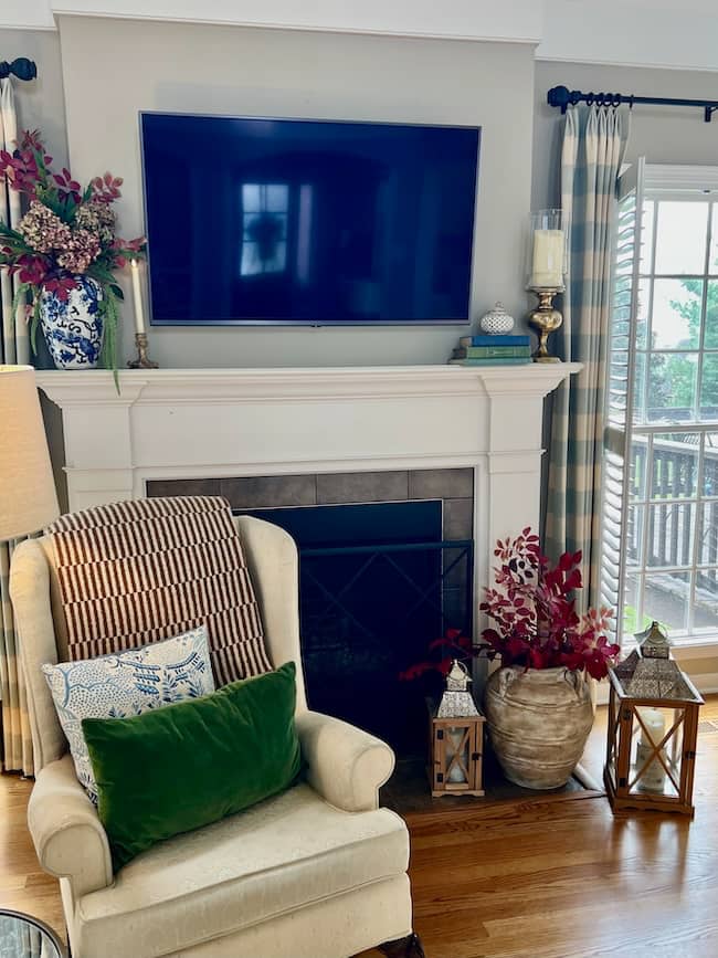 A cozy simple fall home tour scene: a cream armchair with colorful pillows and a green throw sits by a fireplace beneath a TV. Decorative vases, lanterns, and flowers adorn the mantel and floor, while curtains frame the glass doors.
