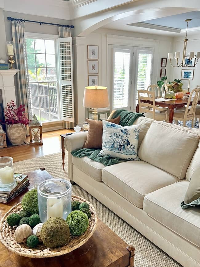Bright living room with a cream sofa, blue and white pillow, and a wooden coffee table topped with moss balls and candles—perfect for a cozy simple fall home tour. In the background, a dining area with large windows lets in natural light.