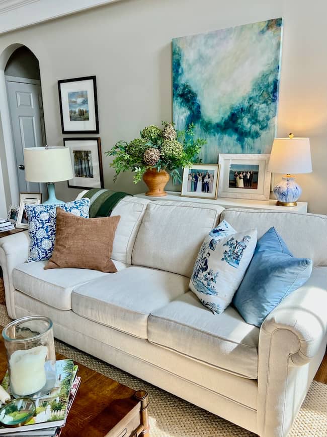 A cozy simple fall home tour features a cream-colored sofa with blue and brown pillows, a wooden coffee table, framed art and photos, a table lamp, and a vase of flowers on the console table against a warm beige wall.
