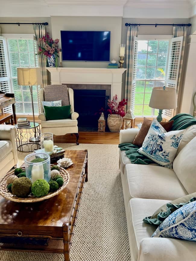 A cozy living room perfect for a simple fall home tour, featuring a white sofa, patterned pillows, a wooden coffee table with candles and moss decor, an armchair, and a fireplace under a wall-mounted TV. Large windows let in natural light and frame the greenery outside.