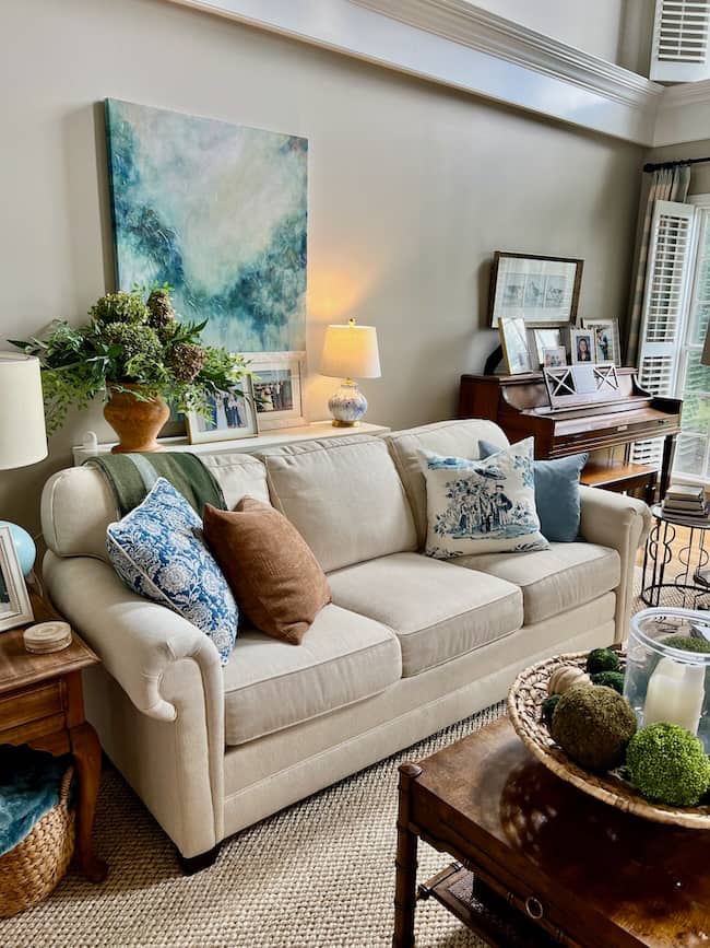 A cozy, simple living room features a beige sofa with decorative pillows, a side table with a lamp, a large abstract painting, and a piano holding framed photos—perfect for your fall home tour with natural light streaming through nearby windows.