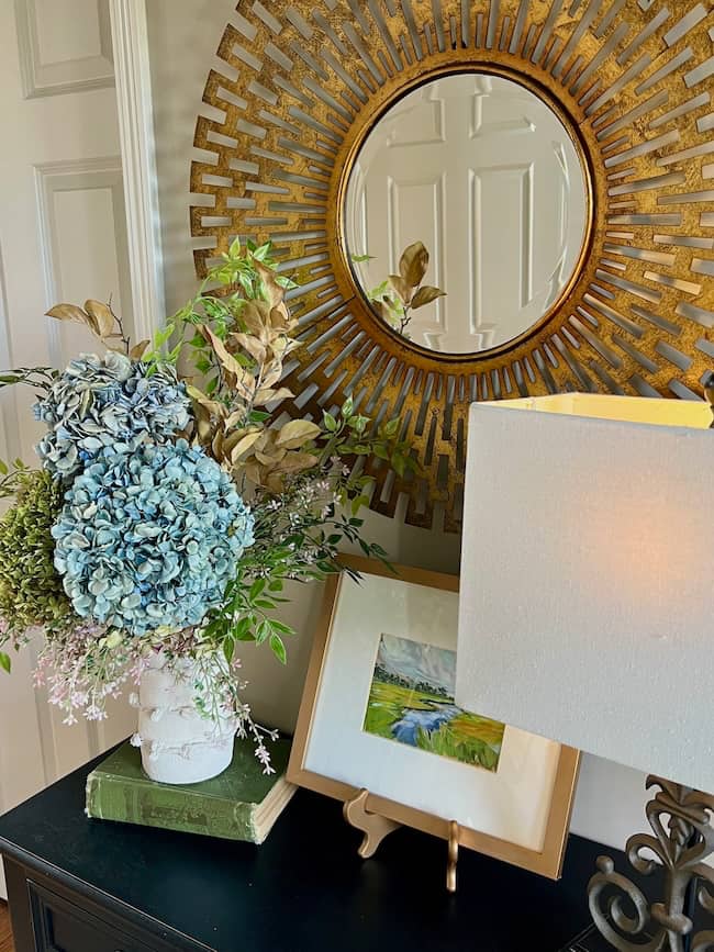 A decorative table holds a vase of blue hydrangeas and foliage, a green book, a framed landscape painting, and a lamp with a white shade—perfect touches for a cozy simple fall home tour. A round, gold sunburst mirror hangs above.