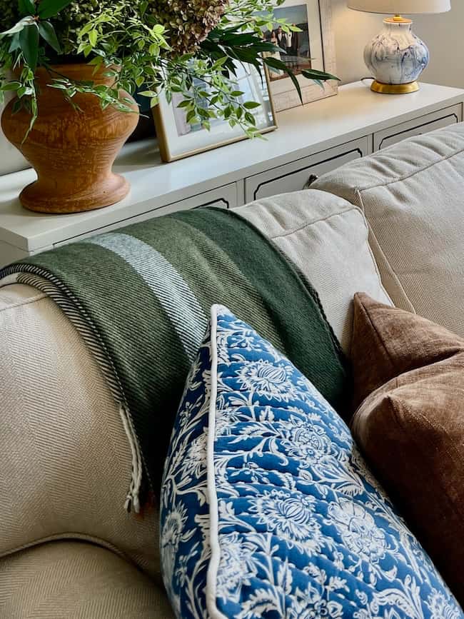 A beige sofa with a green striped throw, blue and white floral pillow, and brown velvet pillow creates a cozy vibe. Behind the sofa, a white console with a potted plant, framed photos, and lamp completes this simple fall home tour look.
