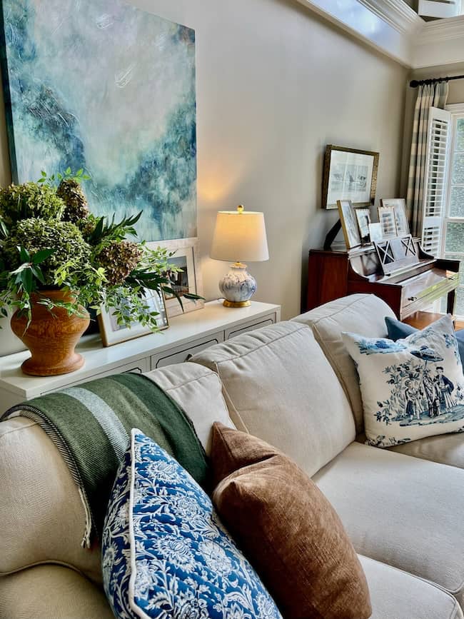 A cozy living room perfect for a simple fall home tour, featuring a beige sofa with blue, brown, and green pillows, a green throw, large abstract painting, potted plant, table lamp, framed photos, and natural light from nearby windows.