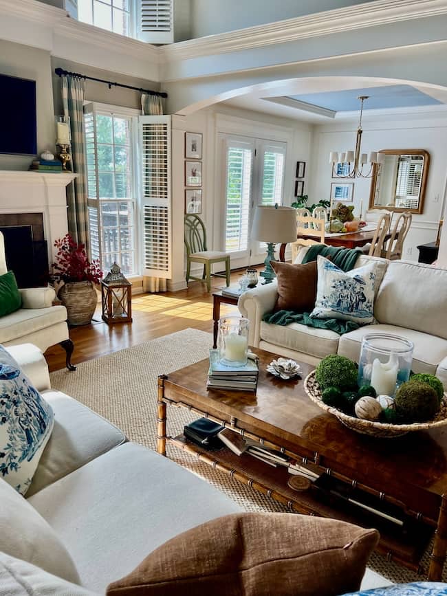 Bright, airy living room with large windows, white sofas with patterned pillows, and a wooden coffee table—perfect for a cozy simple fall home tour. Enjoy an open view into the dining area set for a meal beneath a sparkling chandelier.