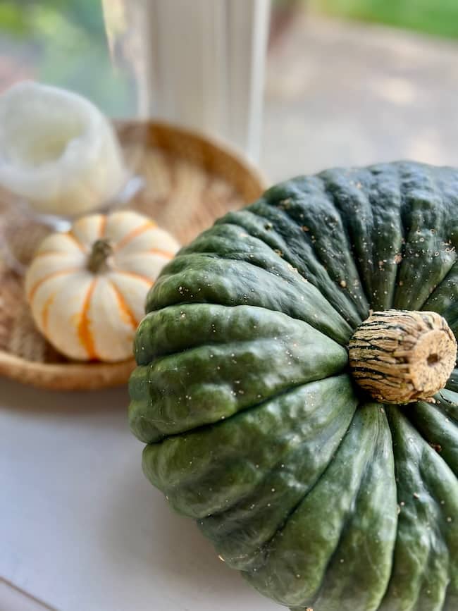 A large, dark green pumpkin sits on a white surface next to a small white pumpkin with orange stripes and a white candle on a woven tray, perfect for a cozy simple fall home tour by a bright window.
