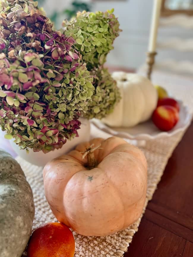 A cozy simple fall home tour centerpiece features a peach pumpkin, white pumpkin, dried hydrangea flowers in a vase, apples, and an orange on a textured beige runner, with a candlestick visible in the background.