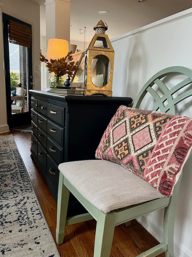 A green chair with a red and pink patterned pillow sits next to a black dresser topped with cozy fall decor—a lamp, gold lantern, vase of dried flowers, and mirror—in a well-lit hallway with wooden floors, perfect for a simple fall home tour.