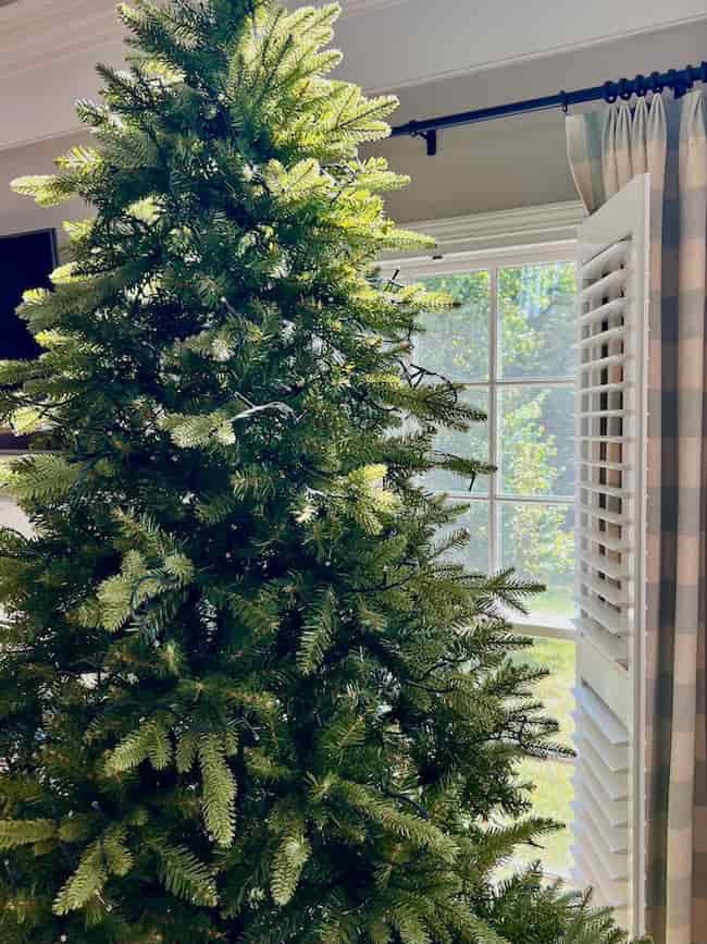 A green artificial Christmas tree stands indoors near a window with open white shutters, filling the bright room with natural light—perfect for learning how to fluff and light an artificial Christmas tree before decorating.