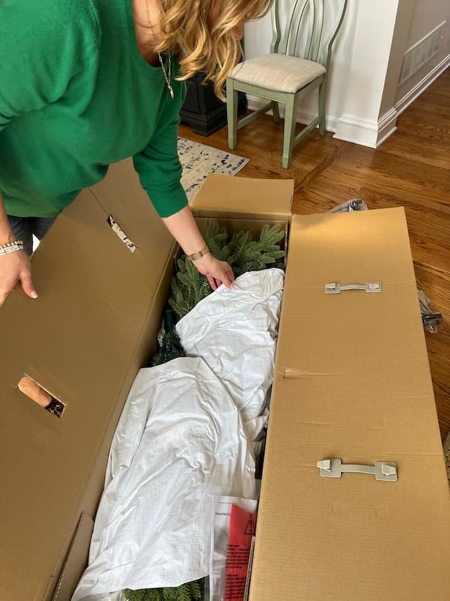 A person in a green sweater is opening a large cardboard box filled with artificial greenery, possibly a Christmas tree, covered with white fabric—perhaps getting ready to show how to fluff and light an artificial Christmas tree. A rug and chair are in the background.
