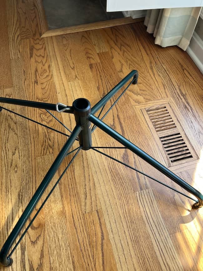 A metal Christmas tree stand with four legs sits on a hardwood floor near a vent and a light-colored curtain, bathed in sunlight—the perfect spot for learning how to fluff and light an artificial Christmas tree.