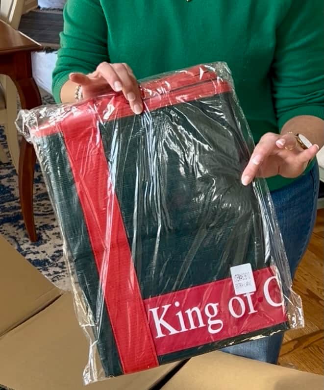 A person in a green sweater holds a packaged red and green item—possibly decorations or instructions on how to fluff and light an artificial Christmas tree—with "King of C" partially visible on the packaging, still wrapped in clear plastic.