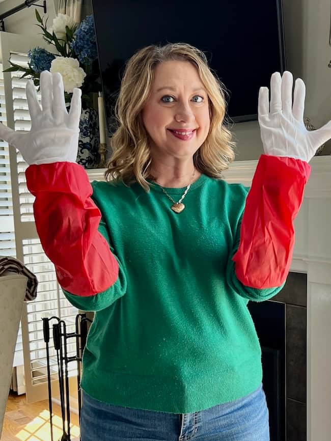 A smiling woman in a green sweater and blue jeans shows off red and white Santa gloves, standing indoors near a fireplace and window with plants—perhaps ready to demonstrate how to fluff and light an artificial Christmas tree.