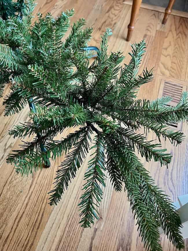 Artificial pine tree branches in a stand, viewed from above, set on a wooden floor with part of a chair visible—an ideal setup for learning how to fluff and light an artificial Christmas tree beautifully.