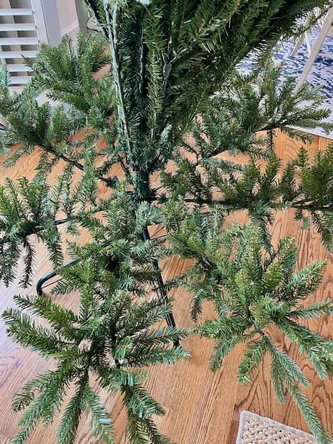 A partially assembled artificial Christmas tree stands on a wooden floor, its branches spread out near the base as if ready for the next steps in how to fluff and light an artificial Christmas tree. The upper section remains unfluffed and undecorated.