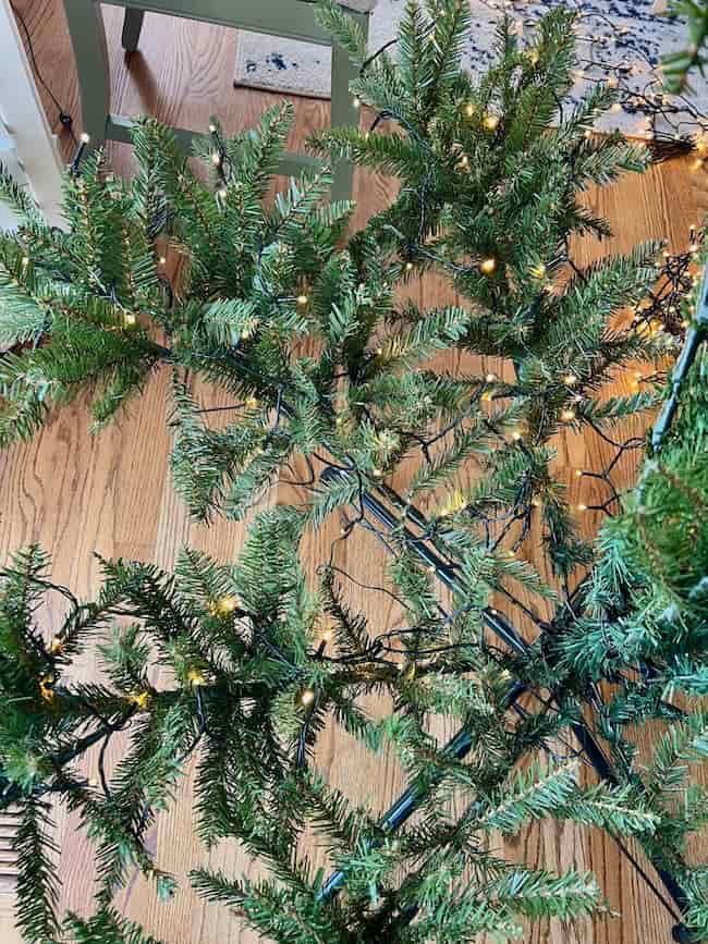 A partially assembled artificial Christmas tree with green branches and lights sits on a wooden floor, viewed from above—perfect for learning how to fluff and light an artificial Christmas tree.