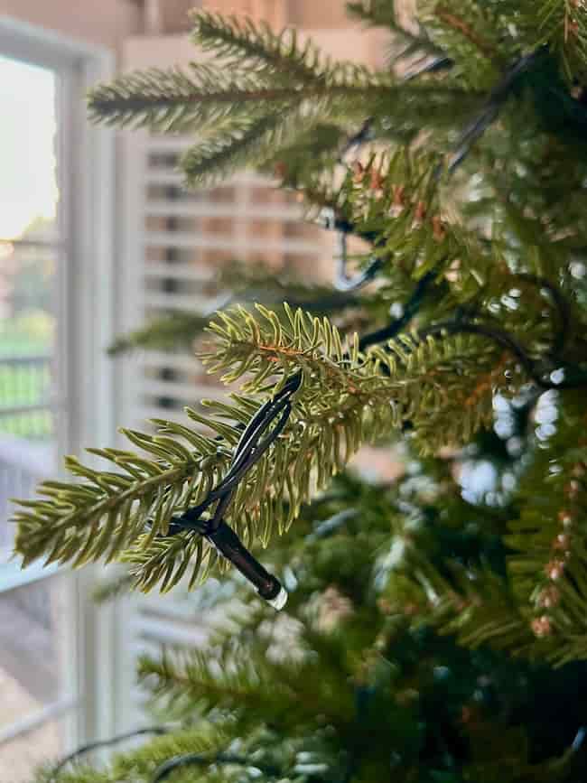 Close-up of artificial pine tree branches wrapped with black string lights, showcasing how to fluff and light an artificial Christmas tree near a window with daylight softly illuminating the blurred background.