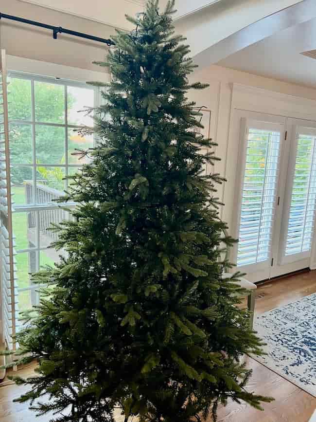 A large, undecorated artificial Christmas tree stands in a bright living room near glass doors and windows with white shutters. Sunlight filters in, creating the perfect spot for learning how to fluff and light an artificial Christmas tree.