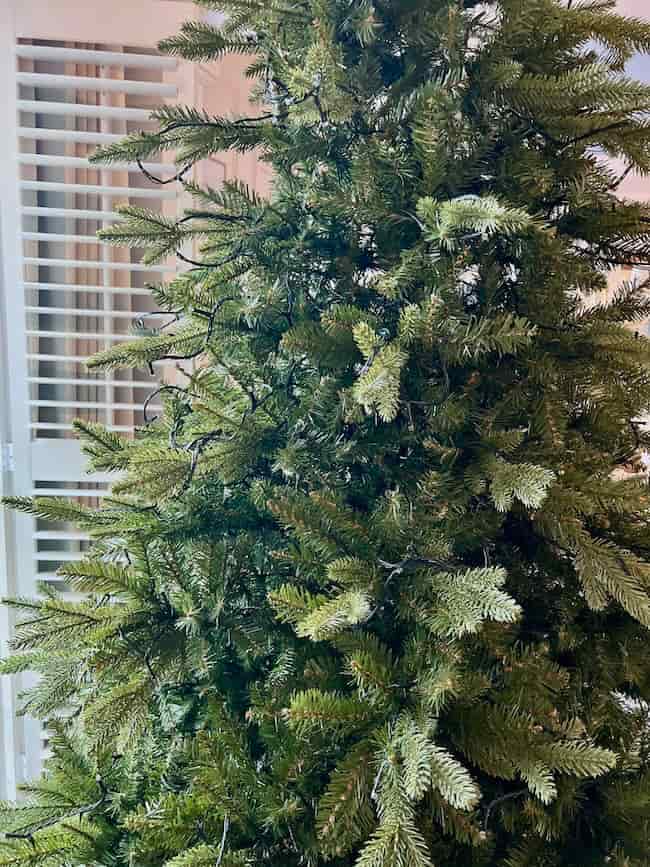 A close-up of an undecorated artificial Christmas tree with string lights, standing indoors near a window with white blinds—perfect for showing how to fluff and light an artificial Christmas tree.
