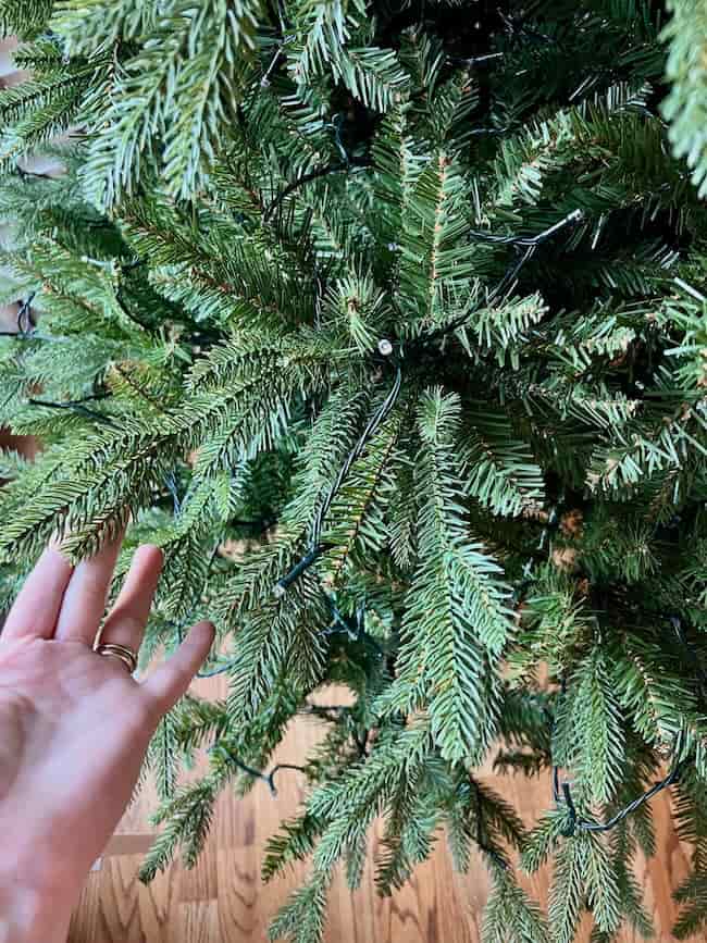A hand reaches toward the branches of an artificial Christmas tree decorated with a string of lights, showing how to fluff and light an artificial Christmas tree, set on a wooden floor.