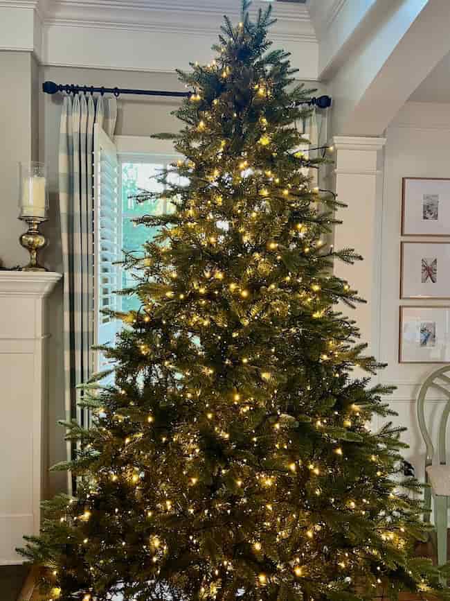 A Christmas tree, perfectly fluffed and lit with warm white string lights, stands in a living room near a window with striped curtains and framed artwork. Want tips? Learn how to fluff and light an artificial Christmas tree for a cozy holiday look.