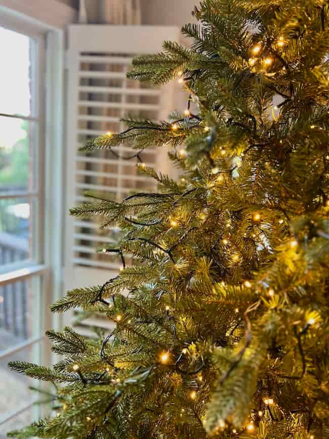 A close-up of a Christmas tree decorated with warm white string lights, set near a sunlit window with white blinds in the background—perfect inspiration for how to fluff and light an artificial Christmas tree beautifully.