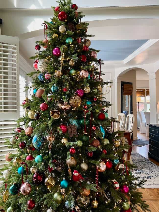 A large Christmas tree, perfectly fluffed and lit as per how to fluff and light an artificial Christmas tree tips, is adorned with colorful ornaments—red, gold, blue, and green baubles—in a bright living room with white walls and classic furnishings.