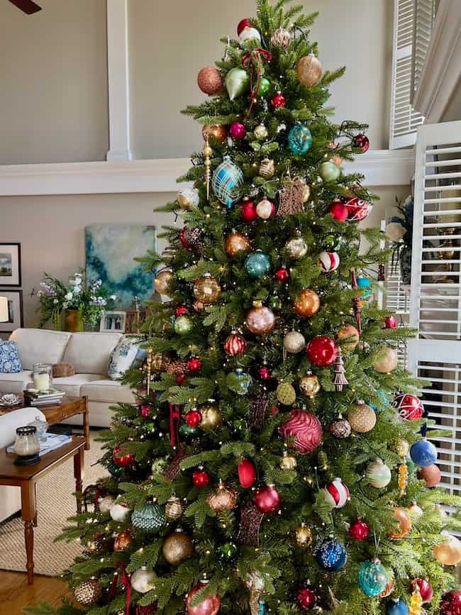 A tall, lush Christmas tree—expertly fluffed and lit following tips on how to fluff and light an artificial Christmas tree—stands decorated with colorful ornaments in a sunlit living room with white shutters.