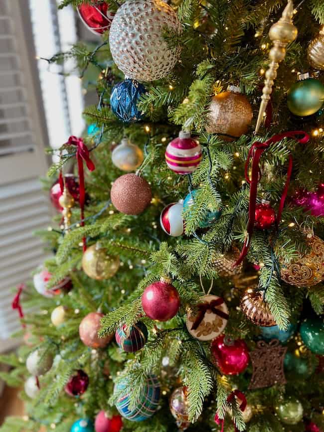 A decorated Christmas tree with colorful ornaments, including red, gold, blue, and striped balls, and ribbon bows, shows how to fluff and light an artificial Christmas tree, with white lights glowing among the green branches.