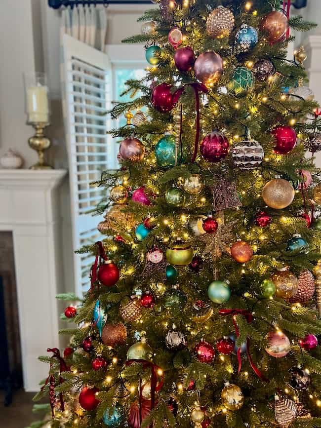 A Christmas tree, expertly fluffed and brightly lit using tips on how to fluff and light an artificial Christmas tree, stands near a white fireplace and a window with shutters adorned in vibrant ornaments.