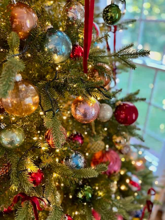 A Christmas tree decorated with colorful ornaments and twinkling lights stands near a sunlit window, creating a festive and cozy holiday atmosphere—showing the perfect result of knowing how to fluff and light an artificial Christmas tree.