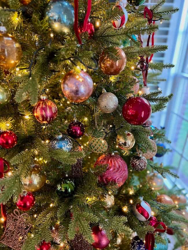 A Christmas tree, expertly fluffed and lit, is decorated with colorful ornaments—red, gold, and green baubles—and glows with warm white string lights near a bright window. Learn how to fluff and light an artificial Christmas tree for a festive look.