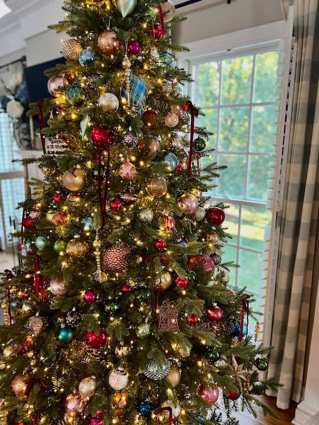 A decorated Christmas tree, expertly fluffed and lit, stands by a large window, adorned with multicolored ornaments and ribbon in a cozy, sunlit room with plaid curtains.