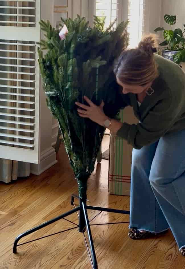 A woman demonstrates how to fluff and light an artificial Christmas tree, adjusting the bundled branches in a bright room with wood floors and plants in the background.