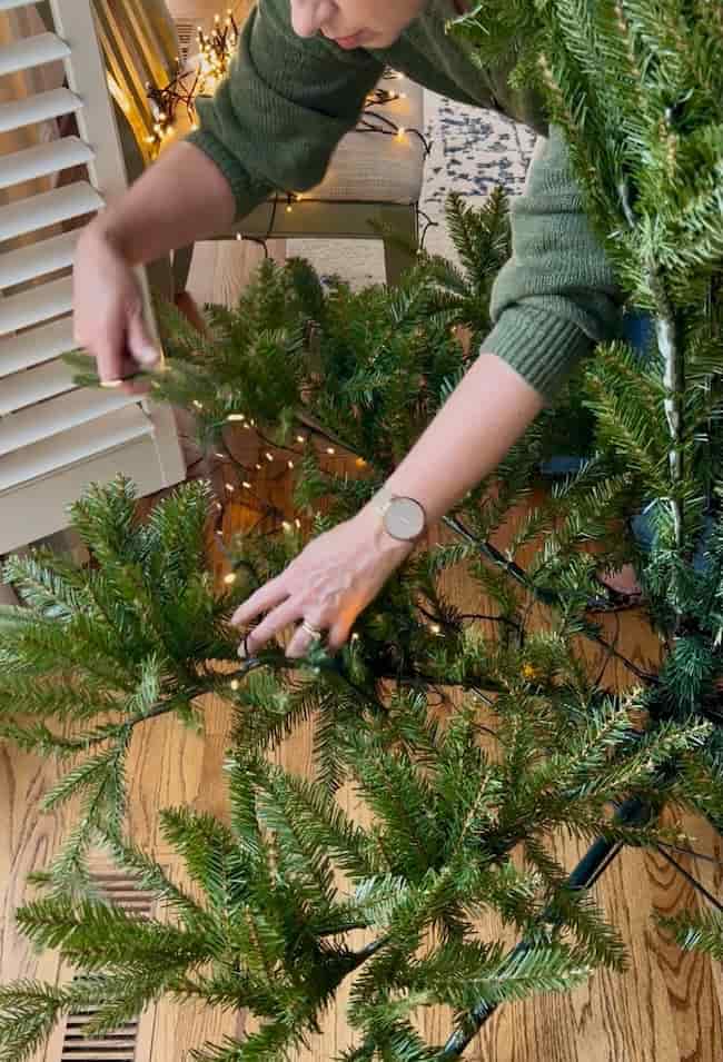 A person in a green sweater demonstrates how to fluff and light an artificial Christmas tree, adjusting its branches while string lights twinkle indoors on a wooden floor.