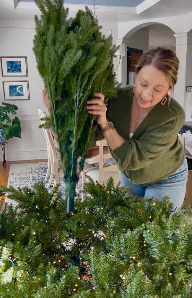 A woman in a green sweater sets up the top section of an artificial Christmas tree in a living room, showing how to fluff and light an artificial Christmas tree, with string lights already on the branches.