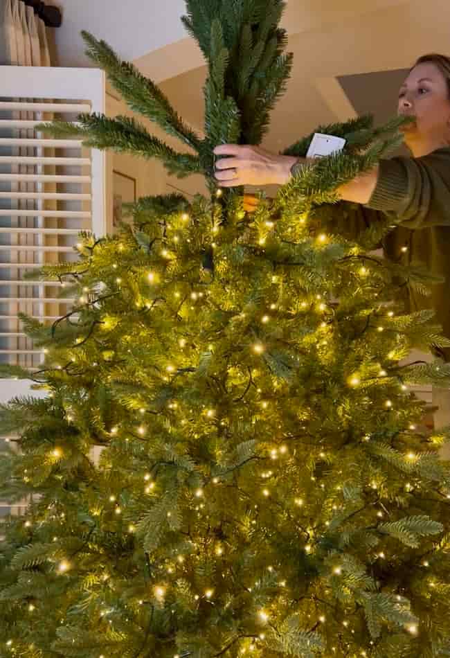 A woman stands beside a Christmas tree decorated with warm white string lights, demonstrating how to fluff and light an artificial Christmas tree in a cozy indoor setting.