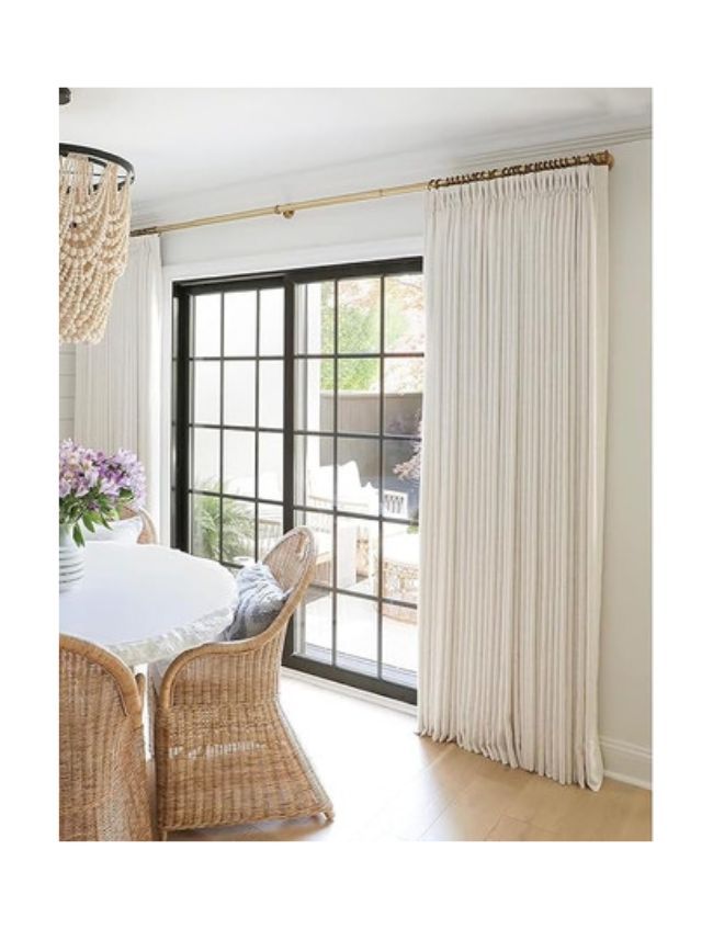 A bright dining area with rattan chairs and a round table topped with purple flowers, next to floor-to-ceiling cream curtains and large black-framed sliding glass doors, seamlessly complementing classic cozy living room decor.
