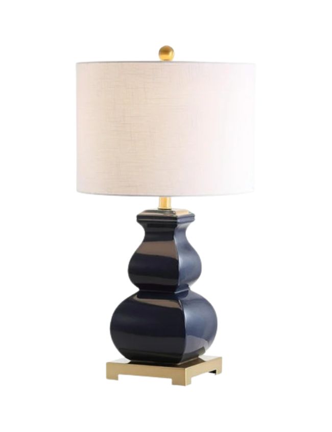 A table lamp with a glossy dark blue, curvy ceramic base, gold finial and accents, and a large white cylindrical fabric lampshade. Its stylish design makes it perfect for classic cozy living room decor.