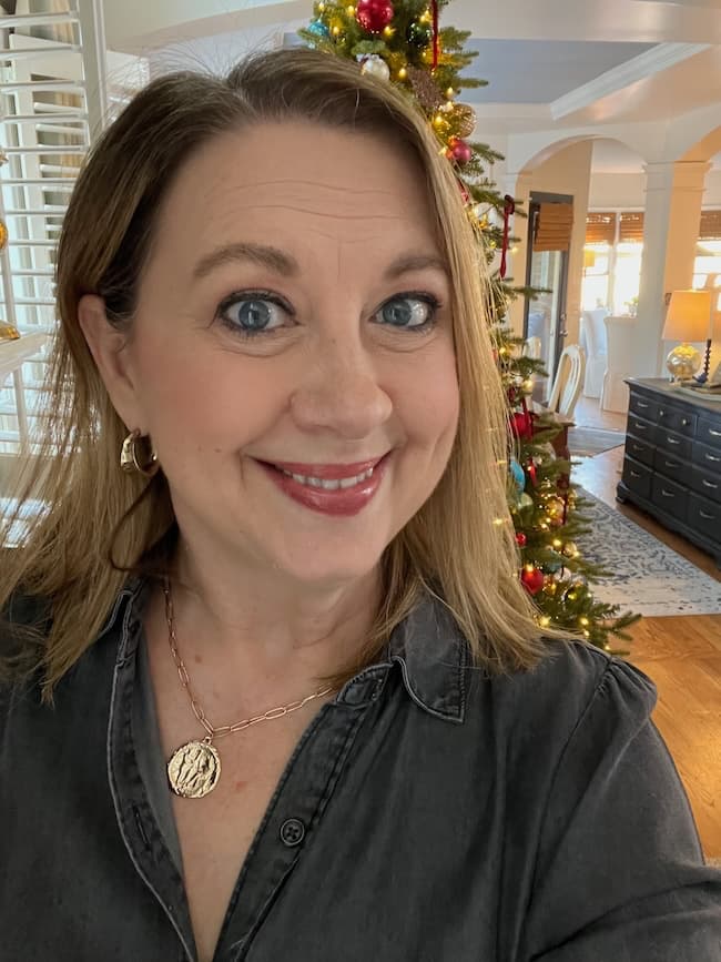 A woman with light brown hair smiles for a selfie indoors, standing by a decorated Christmas tree. She wears a dark shirt, gold earrings, and a necklace, radiating cozy weekend vibes in her warmly lit living room.