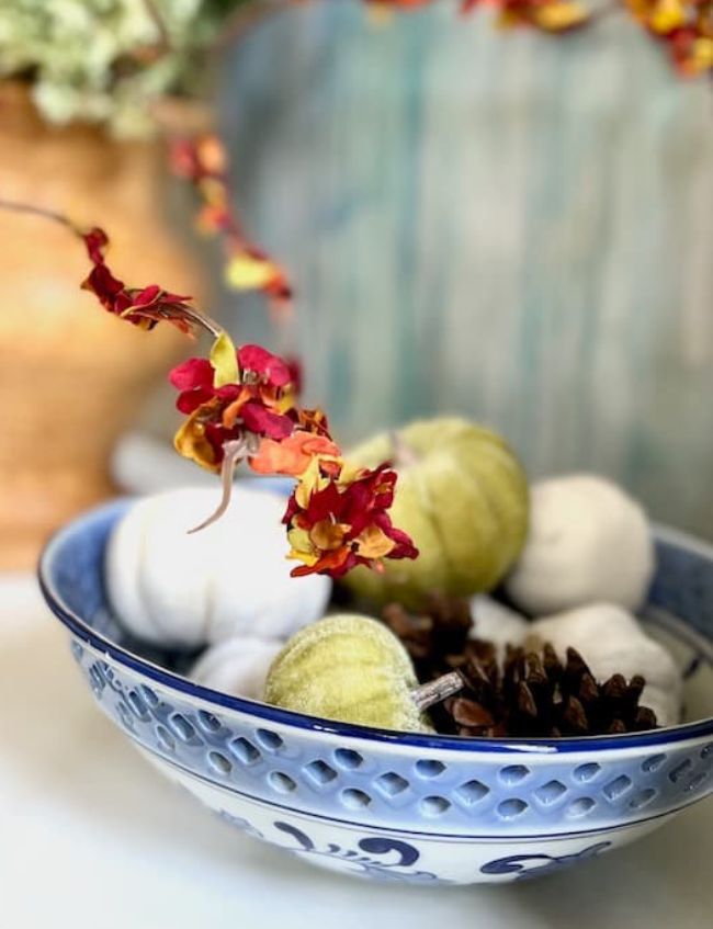 A decorative blue and white bowl holds small white and green pumpkins and pinecones, with an orange and red floral branch arching above. The softly blurred background adds a warm and cozy touch, perfect for weekend inspiration.