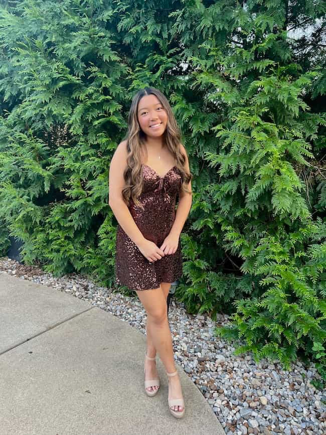 A young woman with long hair stands outdoors on a sidewalk, smiling. She is wearing a sparkly dark brown dress and light-colored heels, with green bushes in the background—true Weekend Inspiration from Seven on Saturday.