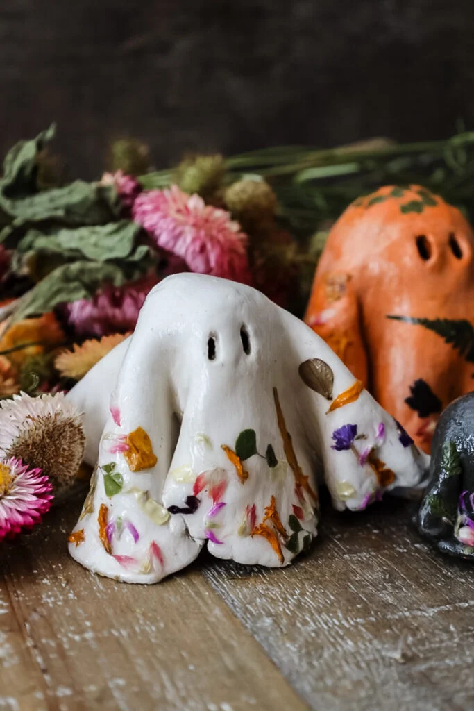 A small ghost-shaped figure with a white surface decorated in colorful dried flower petals sits on a wooden surface, surrounded by other Cozy Start ghost figures and dried blooms—capturing the spirit of the season.