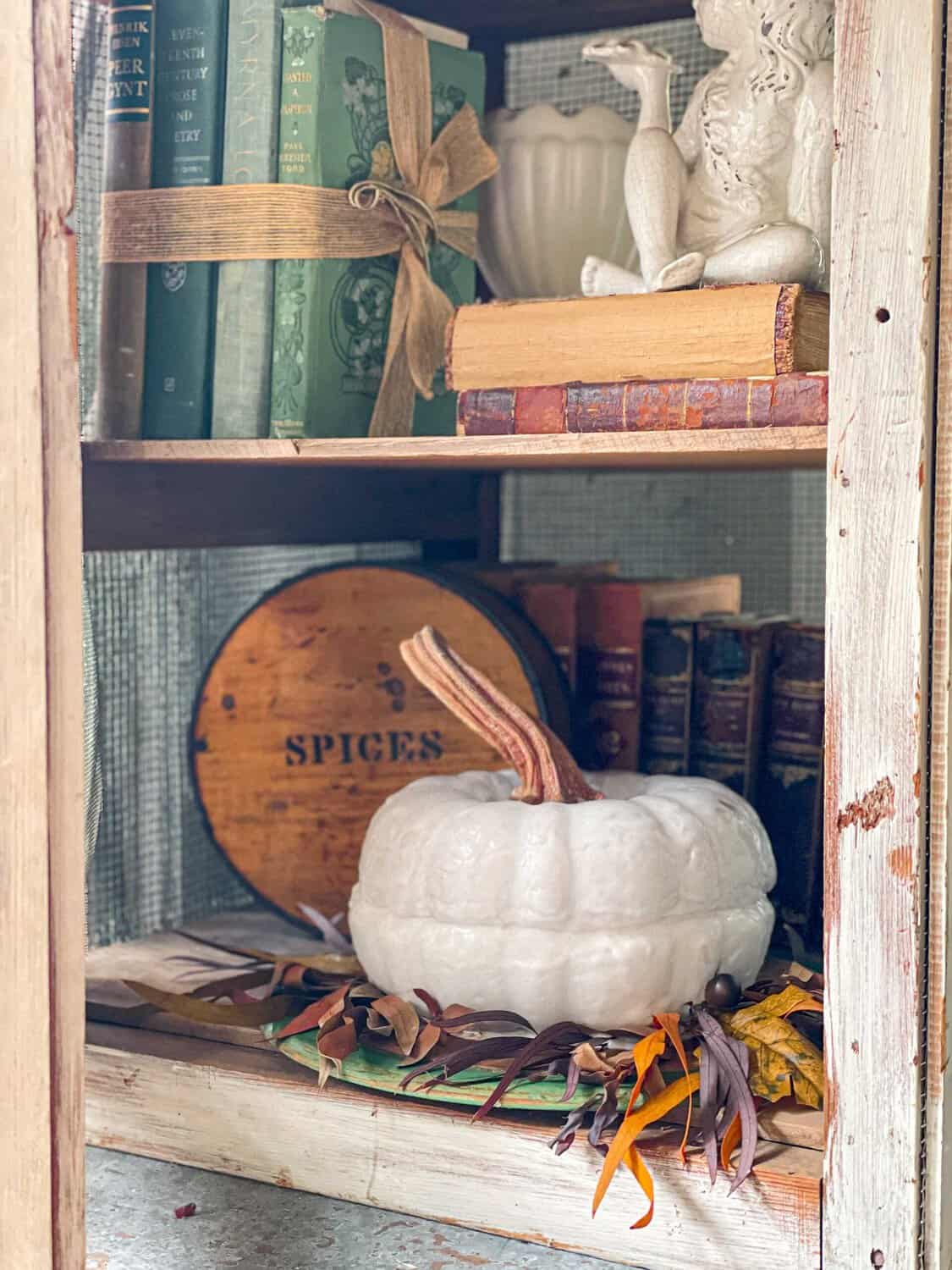A rustic bookshelf offers fresh inspiration with vintage books, a wooden "Spices" box, a white pumpkin surrounded by autumn leaves, and a white decorative dragon figurine—perfect for cozy October décor.