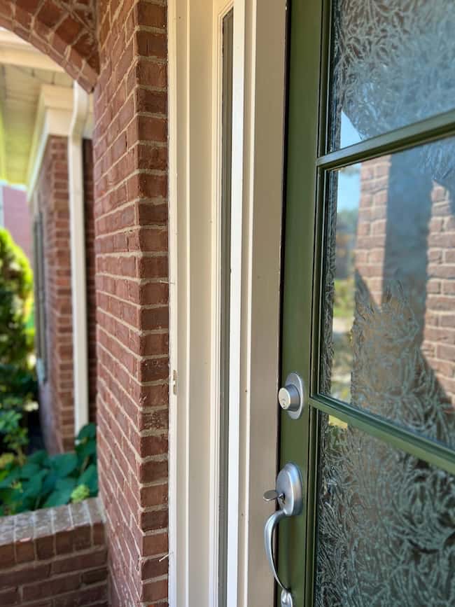 Close-up of a dark green front door with frosted glass panels and a silver handle on a brick house, showcasing a classic look, with part of the porch and greenery visible to the left.
