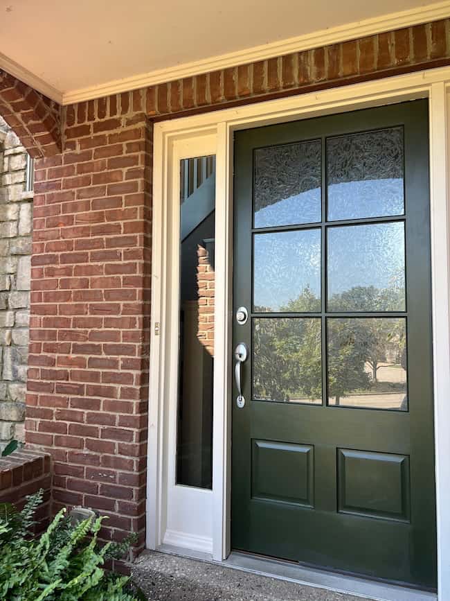 A dark green front door with glass panels and a silver handle is set in a brick wall, offering a classic look. A tall, narrow window sits to the left, with plants at the bottom corner and trees visible through the glass.