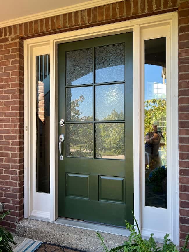 A dark green front door with four glass panels and silver hardware offers a classic look, framed by two narrow vertical windows in a brick exterior. Trees and a person’s reflection appear in the glass.